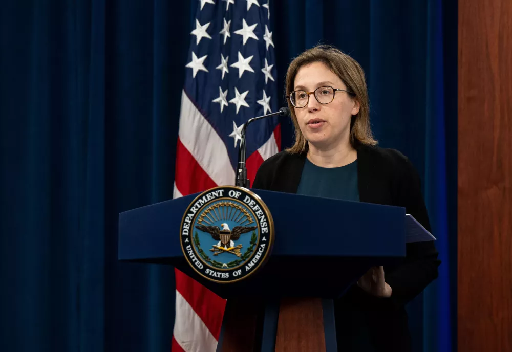 Sasha baker speaking at a podium with the seal of the Department of Defense. In the background, an American flag.