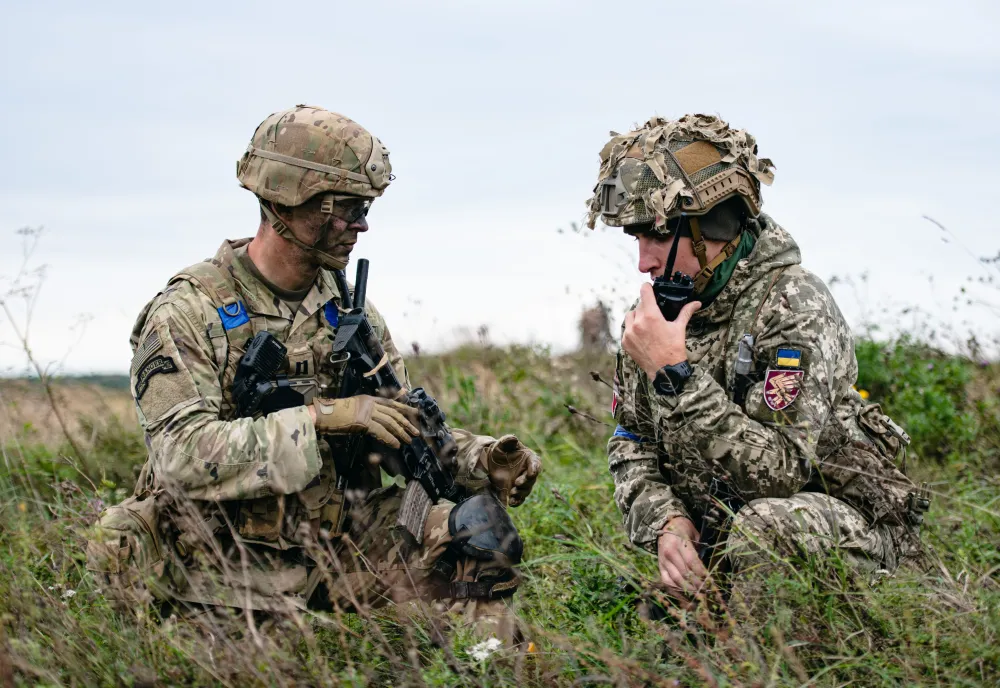 An American paratrooper and a Ukrainian paratrooper kneel next to each other on a drop zone.