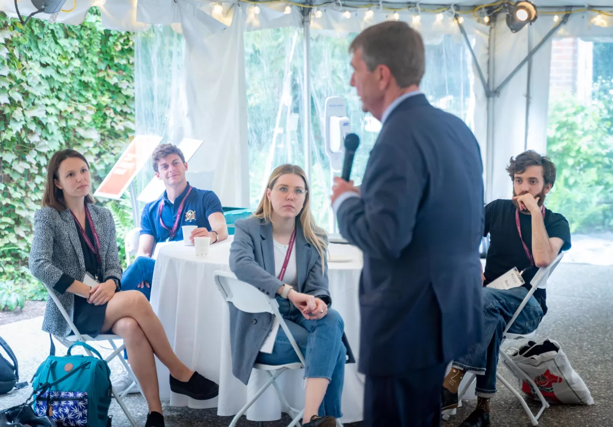 Ash Carter in the foreground with a microphone speaking to students, in focus in the background, seated around a table.