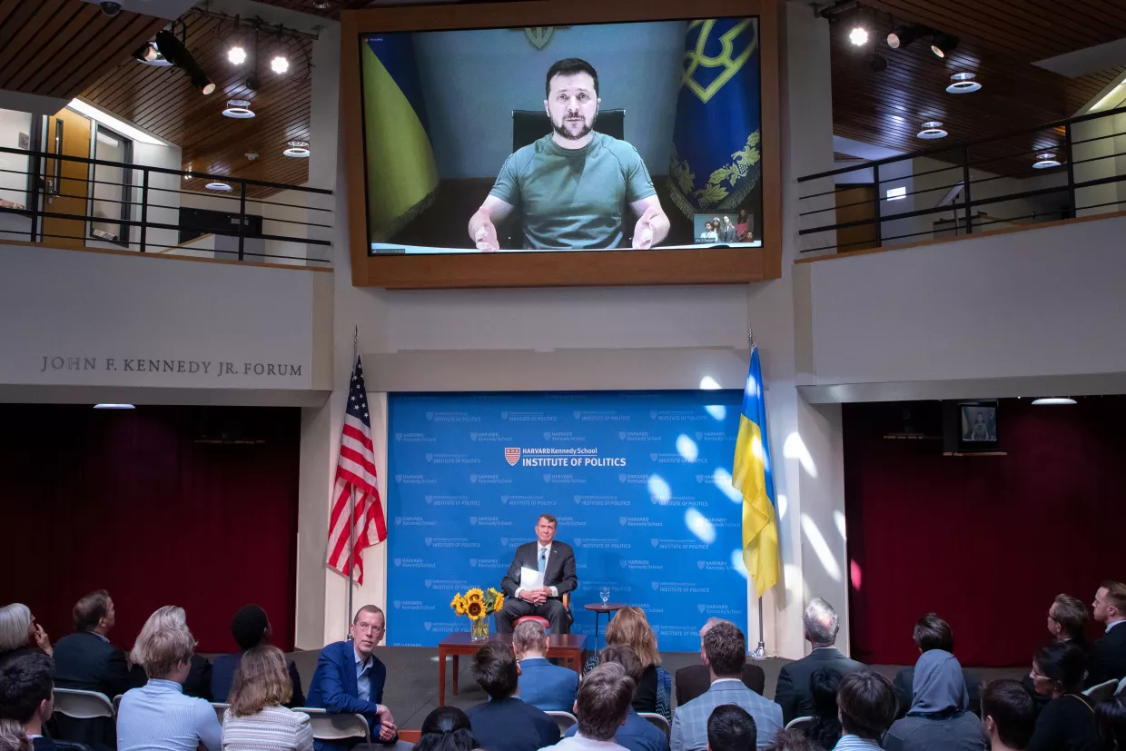 Ukrainian President Zelenskyy on the screen above Ash Carter, sitting on the stage in the JFK Jr. Forum with a crowd of people, including HKS Dean Doug Elmendorf in the foreground.