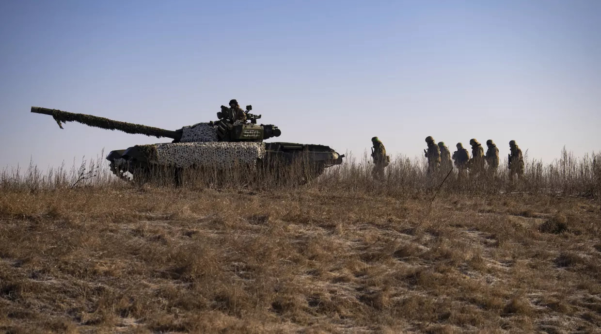 A tank rolls across a dry field with soldiers walking behind it.