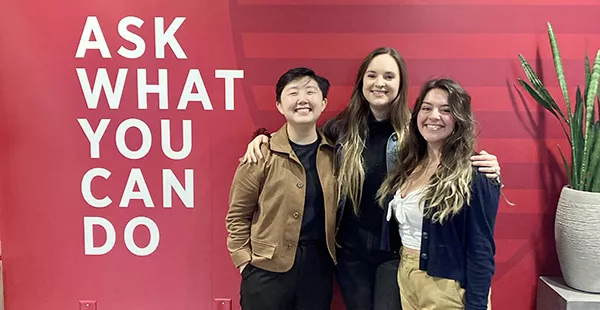 three students smile and pose next to a sign that says "Ask what you can do"