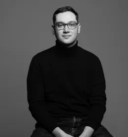 Black and white photo of a man sitting on a stool facing the camera.