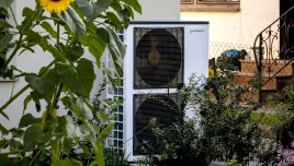 A heat pump flanked by sunflowers against the side of a house.