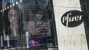 In this Nov. 9, 2020, file photo, an ad for COVID-19 testing reflects on glass at a bus stop, as pedestrians walk past Pfizer world headquarters in New York. 