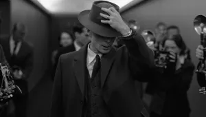 A black and white image from the film Oppenheimer showing Cillian Murray as Oppenheimer with his hat puled down over his face shielding himself from press photographers.