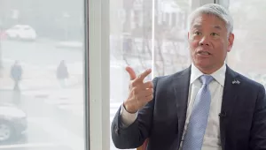 John Tien speaking in front of a window with Harvard Square in the background