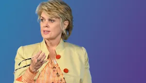 A woman with cropped blonde hair in a yellow jacket gestures while speaking.