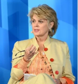 A woman with cropped blonde hair in a yellow jacket gestures while speaking.