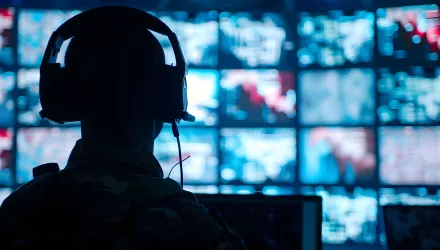 Silhouette of a military man in headphones at a laptop against the background of a video wall of glowing screens 