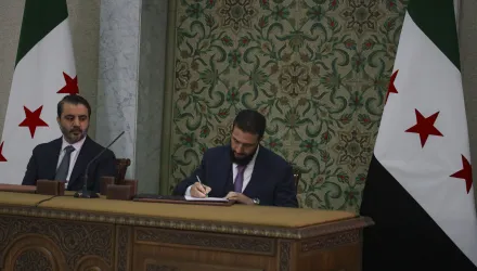 Ahmad Al-Sharaa signs a temporary constitution while next to Asaad Hassan al-Shiban, seated at a desk alongside two Syrian flags