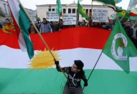 A Kurdish protester poses with a small Kurdish flag in front of a large Kurdish flag.