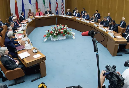 U.S. and Iranian negotiators sit around a cured table as media with cameras and microphones crowd the foreground.