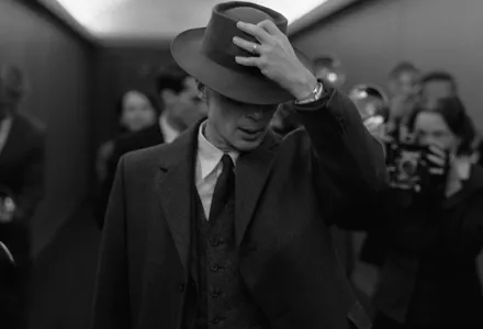 A black and white image from the film Oppenheimer showing Cillian Murray as Oppenheimer with his hat puled down over his face shielding himself from press photographers.