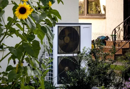 A heat pump flanked by sunflowers against the side of a house.