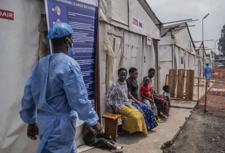 A healthcare worker in protective gear walks past people sitting on a bench outside awaiting treatment for mpox.