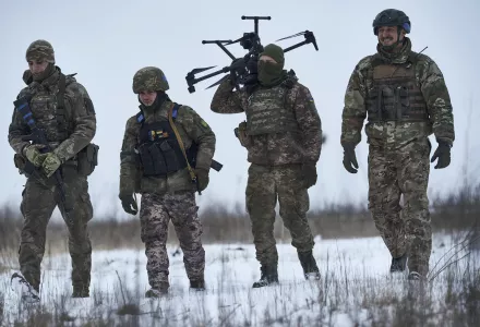 Four Ukrainian soldiers carry a drone as they walk across a snow-covered field. 