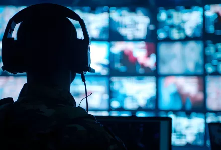 Silhouette of a military man in headphones at a laptop against the background of a video wall of glowing screens 