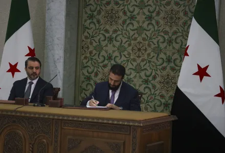 Ahmad Al-Sharaa signs a temporary constitution while next to Asaad Hassan al-Shiban, seated at a desk alongside two Syrian flags